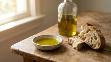 A shallow ceramic dish of golden-green olive oil beside torn chunks of crusty sourdough on a rustic wooden table