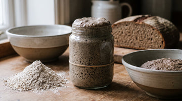 A jar of active rye sourdough starter beside rye flour and a rye loaf